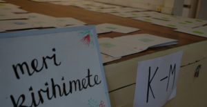 photograph of a Holiday Card Drive, with whiteboard reading "Meri Kirihimete" in front of a stage covered in large envelopes.